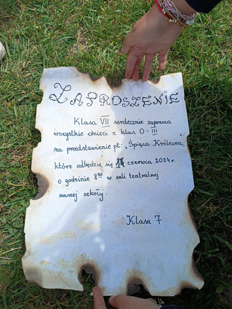 Na trawniku ułożona jest przypalona na brzegach kartka papieru z odręcznym napisem: "ZAPROSZENIE Klasa VII serdecznie zaprasza wszystkie dzieci z klas 0 - III na przedstawienie pt: "Śpiąca Królewna", które odbędzie się 17 czerwca 2024 r. o godz. 8.50 w sali teatralnej naszej szkoły. Klasa 7".