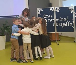 Uczniowie oddziału przedszkolnego przytulają się do swojej wychowawczyni. Z boku stoi tablica z białym napisem "Czas rozwinąć skrzydła".