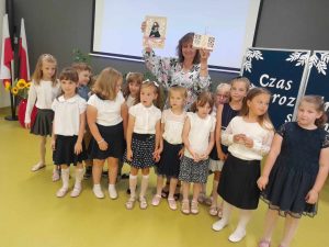 Wychowawczyni oddziału przedszkolnego pozuje do wspólnego zdjęcia wraz ze swoimi uczniami, którzy ją otaczają. Pani podnosi w górę dwa prezenty, które otrzymała od swoim podopiecznych.