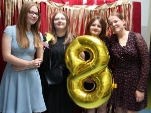 Cztery uczennice pozują do wspólnego zdjęcia. Jedna z nich trzyma złoty balon w kształcie cyfry 8 a druga, kartonik z grafiką pucharu. Dziewczyny pozują na tle czerwonej ściany ozdobionej złotymi paskami.