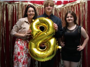 Doje uczniów pozuje do wspólnego zdjęcia ze swoją nauczycielką. Uczeń stojący po środku oraz nauczycielka trzymają przed sobą złoty balon w kształcie cyfry 8. Zdjęcie wykonane na tle czerwonego materiału ozdobionego złotymi paskami.