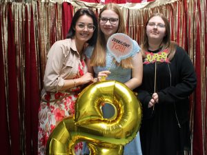 Dwie uczennice pozują do wspólnego zdjęcia ze swoją nauczycielką. Dziewczyna stojąca po środku przytula się do nauczycielki trzymając w dłoni złoty balon w kształcie cyfry 8 i tabliczkę z napisem "ZROBILIŚMY TO". Uczennica stojąca obok trzyma w dłoni tabliczkę z napisem "SUPER". Zdjęcie wykonane na czerwonym tle ozdobionym złotymi paskami.