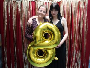 Dwie uczennice klasy ósmej pozują do wspólnego zdjęcia podczas balu. Jedna z dziewcząt obejmuje koleżankę.. Dziewczynki trzymają w dłoniach przed sobą złoty balon w kształcie cyfry 8 i stoją na czerwonym tle ozdobionym złotymi wstążkami.