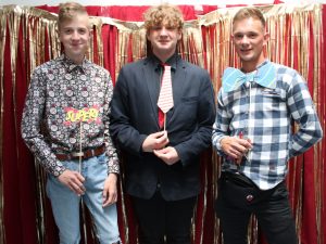 Dwóch uczniów klasy ósmej pozuje do wspólnego zdjęcia z pracownikiem szkoły. Każdy z nich trzyma przed sobą tabliczkę w innym kształcie. Zdjęcie wykonane na czerwonym tle ozdobionym złotymi paskami.