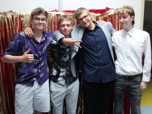 Czworo uczniów klas8 pozuje do wspólnego zdjęcia. Chłopcy obejmują się przedramionami. Jeden z nich ma rozstawione palce w dłoni a drugi podnosi w górę kciuk. W tle widać czerwony materiał ozdobiony złotymi wstążkami.