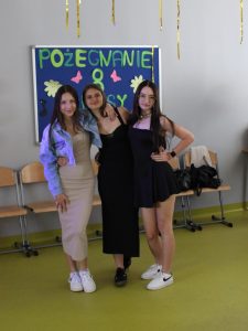Trzy uczennice biorące udział w balu pozują do wspólnego zdjęcia obejmując się. W tle widoczna jest tabliczka z kolorowym napisem "POŻEGNANIE 8 KLASY".