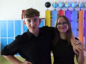 Dwoje uczniów pozuje do wspólnego zdjęcia. Chłopak obejmuje koleżankę ramieniem, a ta trzyma jego dłoń w nadgarsku.