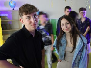 Dwoje uczniów klasy ósmej pozuje do wspólnego zdjęcia podczas balu. Uczniowie stoją w pewnym oddaleniu od siebie a w tle widać rozmazane postacie bawiącej się młodzieży.