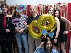 Uczniowie wraz z wychowawczynią pozują do wspólnego zdjęcia. Wychowawczyni trzyma złoty balon w kształcie cyfry 8 oraz tabliczkę z napisem "WOW", którą zasłania twarz ucznia stojącego obok niej. Dwóch uczniów trzyma przed sobą tabliczki z napisami: "KONIEC SZKOŁY" oraz "SUPER", a uczennica kucająca przed nimi trzyma nad głową tabliczkę w kształcie kokardy. Zdjęcie wykonane na czerwonym tle ozdobionym złotymi paskami.