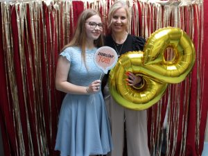 Wychowawczyni klasy pozuje wraz ze swoją uczennicą do wspólnego zdjęcia. Wychowawczyni trzyma przed sobą złoty balon w kształcie cyfry 8, a uczennica tabliczkę z napisem "ZROBILIŚMY TO". Zdjęcie wykonane na czerwonym tle ozdobionym złotymi wstążkami.