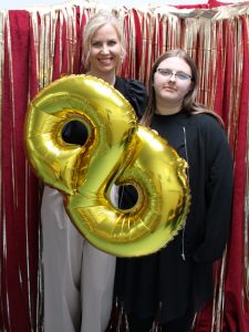 Wychowawczyni klasy pozuje wraz ze swoją uczennicą do wspólnego zdjęcia. Trzymają przed sobą złoty balon w kształcie cyfry 8. Zdjęcie wykonane na czerwonym tle ozdobionym złotymi wstążkami.