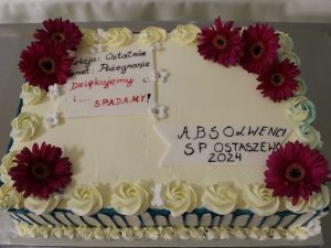 Zdjęcie przygotowanego na bal tortu ozdobionego żywymi kwiatami. Na torcie widnieją napisy: "Lekcja: Ostatnia", "Temat: Pożegnanie", "Dziękujemy i ... SPADAMY", "ABSOLWENCI SP OSTASZEWO 2024".