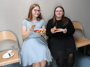 Dwie uczennice klasy ósmej siedzą na krzesełkach na szkolnym korytarzu. Dziewczynki trzymają w dłoniach papierowe talerzyki i kawałki pizzy.