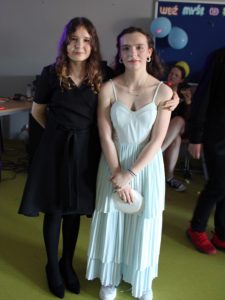 Dwie uczennice klasy ósmej pozują do wspólnego zdjęcia podczas balu. Jedna z dziewcząt obejmuje koleżankę.