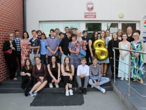 Uczniowie wraz ze swoimi nauczycielami pozują do wspólnego zdjęcia przed budynkiem szkoły. Jedna z uczennic trzyma w dłoni złoty balon w kształcie cyfry 8.