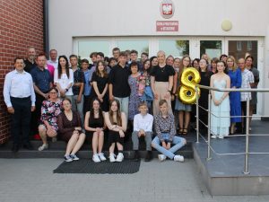 Uczniowie wraz ze swoimi rodzicami i wychowawcami pozują do wspólnego zdjęcia przed budynkiem szkoły. Jedna z uczennic trzyma w dłoni złoty balon w kształcie cyfry 8.
