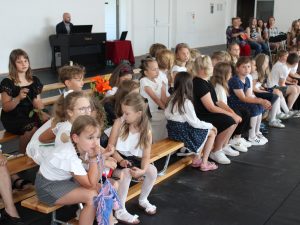 Uczniowie klas 1-3 siedzą na długich drewnianych ławeczkach. Dzieci spoglądają w różne strony.