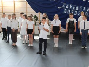 Uczniowie klasy trzeciej stoją w różnych odległościach od siebie na baczność. Z przodu stoi chłopiec trzymający przed sobą sztywną podkładkę z tekstem przemówienia.