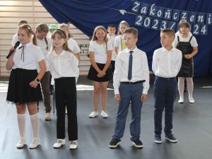 Uczniowie klasy trzeciej stoją podczas wystąpienia. Jedna z dziewczynek trzyma w dłoni mikrofon. Z tyłu stoi uczennica, która szeroko się uśmiecha.