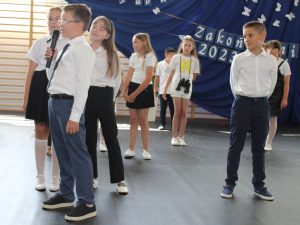 Uczniowie klasy trzeciej stoją podczas wystąpienia na uroczystości. Jeden z chłopców stoi bokiem do pozostałych i przemawia przez mikrofon.