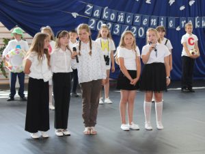Uczniowie klasy trzeciej stoją podczas wystąpienia na uroczystości. Na pierwszym planie stoi w rzędzie pięć uczennic. Dwie z nich trzymają w dłoniach mikrofon.