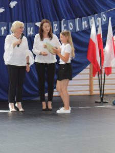 Pani Dyrektor i wychowawczyni klasy trzeciej wręczają uczennicy świadectwo. Dziewczynka szeroko się uśmiecha.
