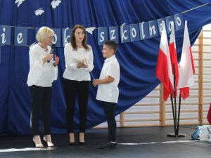 Uczeń klasy trzeciej odbiera świadectwo z rąk swojej wychowawczyni. Na przeciwko ucznia stoi z mikrofonem Pani Dyrektor.