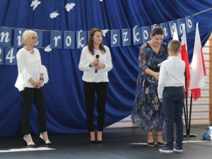Pani bibliotekarka ściska dłoń stojącego na przeciwko ucznia klasy trzeciej. Obok nich stoi Pani Dyrektor i wychowawczyni klasy.
