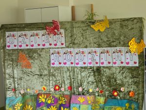 Ma zielonym tle przyczepiony napis: "DZIEŃ RODZINY". Całość ozdobiona kolorowymi motylkami i pracami uczniów z rysunkami kwiatów.