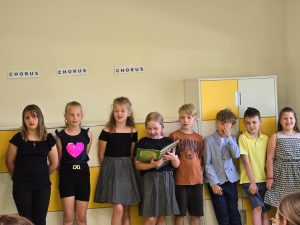 Uczniowie stoją w rzędzie przy ścianie. Jedna z dziewczynek trzyma w dłoniach otwartą książkę, na którą patrzy.