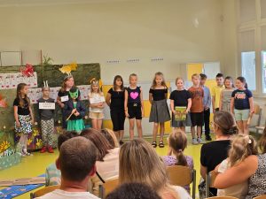 Uczniowie stoją w rzędzie podczas przedstawienia przygotowanego dla rodziców. Jedna z uczennic trzyma w dłoni książkę, a troje innych ma zawieszone na szyi tabliczki z napisami.