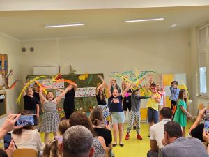 Uczniowie podczas występy stoją po środku sali. Machają wyciągniętymi w górę rękoma, w których trzymają długie, kolorowe wstążki.