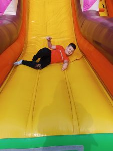 Uczeń zjeżdża z kolorowego dmuchańca. Chłopiec się uśmiecha i leży bokiem.