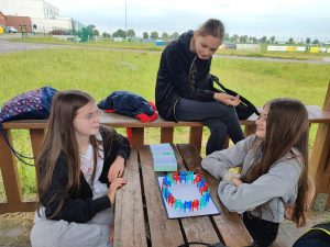 Dwie uczennice siedzą przy stole na którym ułożone są na planszy kolorowe krzesełka. Dziewczynki patrzą na siebie. Na murku za nimi siedzi uczennica przyglądająca się planszy.