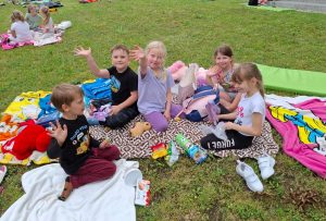 Pięcioro uczniów oddziału przedszkolnego siedzi na kocach na trawniku. Dzieci machają rękoma.