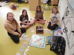Cztery uczennice siedzą na podłodze w sali. Przed nimi leży rozłożona gra monopolu. Dziewczynki patrzą się w stronę obiektywu.