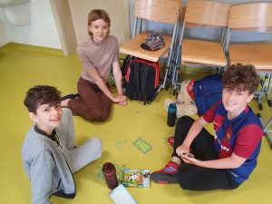 Troje uczniów siedzi na podłodze w sali. Dzieci uśmiechają się do obiektywu. Na podłodze między nimi leży rozłożona gra planszowa.