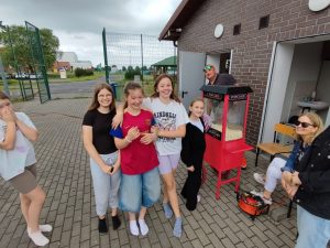 Trzy uczennice uśmiechają się i obejmują. Za nimi widoczna jest maszyna do robienia popcornu przy której stoi nauczyciel i kolejna uśmiechnięta uczennica.