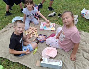 Troje uczniów siedzi na kocu ułożonym na trawie. Pomiędzy nimi leżą drewniane klocki. Dzieci patrzą w stronę obiektywu.