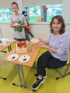 Wychowawczyni zerówki siedzi przy stoliku . W ręku trzyma talerzyk. Przed nią stoi ciasto, które kroi. Obok widoczny jest chłopiec trzymający talerzyk.