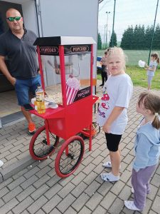 Przy maszynie z popcornem stoi nauczyciel i uczennica. Oboje patrzą się w stronę obiektywu.