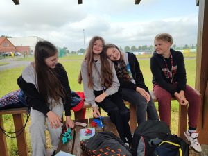 Czworo uczniów siedzi na drewnianej poręczy. Jedna z uczennic opiera głowę o koleżankę siedzącą obok. Wszyscy uczniowie się uśmiechają.