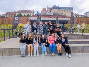 Uczniowie klasy VII wraz z nauczycielką pozują do wspólnego zdjęcia. Uczennice i nauczycielka siedzą na schodach a za nimi stoją chłopcy. W tle widać toruńską starówkę.