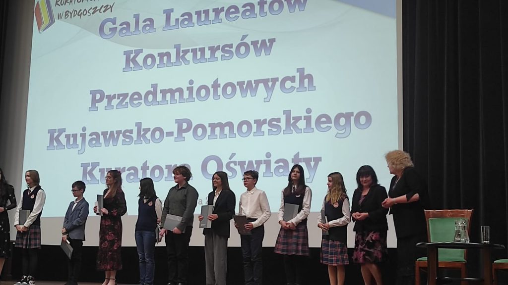 Na tle z wyświetlonym napisem "GALA LAUREATÓW KONKURSÓW PRZEDMIOTOWYCH KUJAWSKO-POMORSKIEGO KURATORA OŚWIATY" stoją w rzędzie uczniowie nagrodzeni w konkursie. Każdy uczeń trzyma przed sobą otrzymany dyplom.