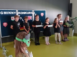 Uczniowie klasy drugiej stoją na tle tablicy z napisem "GMINNY KONKURS POLIGLOTA". Część z uczniów trzyma przed sobą tabliczki z angielskimi wyrazami.