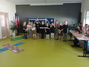Uczniowie klasy drugiej stoją w rzędzie po środku sali. Część z nich trzyma przed sobą tabliczki z angielskimi wyrazami. W tle widać tablicę z napisem "GMINNY KONKURS JĘŻYKOWY". Po prawej stronie widoczne jest siedzące za stołami jury, które nagradza oklaskami występujące dzieci.