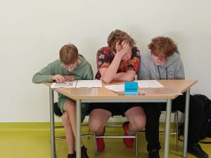 Trzech chłopców siedzi obok siebie przy stole. Wszyscy pochylają się nad kartkami na nim rozłożonymi. Jeden podpiera głowę dłonią.