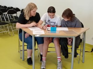 Trzy dziewczynki siedzą przy stole. Wszystkie pochylają się nad kartkami papieru na nim rozłożonymi.