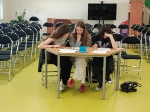 Trzy uczennice siedzą przy wspólnym stole. Wszystkie pochylają się nad kartkami na nim rozłożonymi. Jedna z nich opiera głowę o dłoń dotykającą czoła inna o dłonią podpiera głowę.