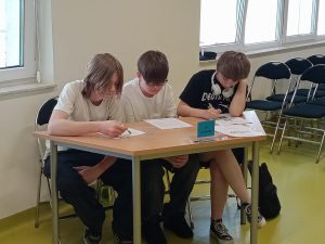 Trzech uczniów siedzi przy wspólnym stole. Każdy z nich patrzy na kartkę papieru leżącą przed nim.